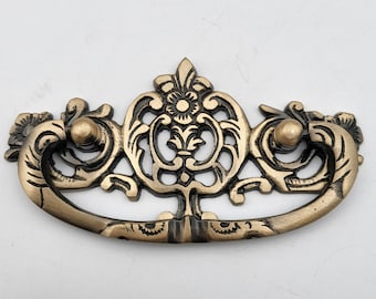 May include: Ornate gold-toned metal drawer pull with a decorative floral design. The pull has a loop shape and is designed for use on cabinets, dressers, and other furniture.