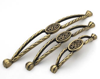May include: Three antique bronze cabinet pulls with an oval, filigree center and twisted handle design. The pulls are in varying sizes, suitable for drawers or cabinets. The hardware has a vintage aesthetic.