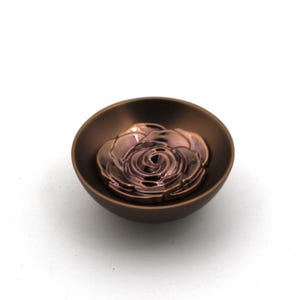 May include: A small, circular, matte brown bowl featuring a metallic, rose-shaped insert at the center. The rose has a polished, reflective finish with deep bronze and purple tones, contrasting with the simple, dark, solid-colored bowl exterior.