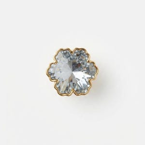 May include: A flower-shaped button featuring a clear, faceted crystal center set within a thin, polished gold-toned metal frame. The design consists of six rounded petals, creating a symmetrical floral silhouette with reflective surfaces.