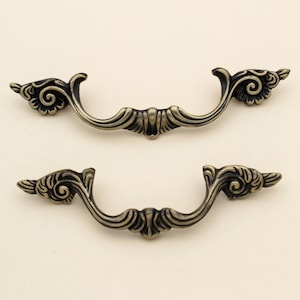 Op de afbeelding: Twee antieke bronzen ladehandgrepen met sierlijke, wervelende bloemmotieven. Elke handgreep heeft een gebogen handvat en decoratieve uiteinden, perfect om een vintage touch aan meubels toe te voegen. De handgrepen staan tegen een neutrale achtergrond.