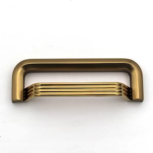 Puede incluir: Un tirador de cajón de metal con acabado en bronce mate en el marco exterior en forma de U y un acabado en oro pulido en la sección interior, que consta de cuatro crestas horizontales, redondeadas y escalonadas a lo largo del ancho.