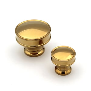 May include: Two gold-toned cabinet knobs with a clear, rounded top and a cylindrical base. The knobs are of different sizes, with a larger one on the left and a smaller one on the right. The knobs are set against a white background.