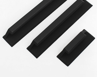 May include: Three black, rectangular cabinet pulls with a ribbed texture. Each pull has a raised handle and a flat base. The pulls are arranged diagonally against a white background, showcasing their design and form.