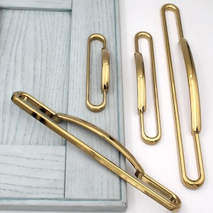 May include: A selection of gold-coloured cabinet pulls and handles in different sizes. The hardware has a sleek, modern design with a curved, rectangular shape. The pulls are displayed against a light blue wooden surface.