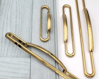 May include: A collection of gold-toned cabinet pulls and handles in various sizes. The hardware features a sleek, modern design with a curved, rectangular shape. The pulls are displayed against a light blue wooden surface.