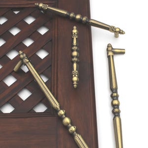 May include: Several antique brass cabinet pulls and handles are displayed against a dark wood door with a lattice design. The hardware features ornate detailing, including decorative finials and a warm, aged finish. The pulls vary in size and shape.