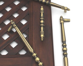 May include: Several antique brass cabinet pulls and handles are displayed against a dark wood door with a lattice design. The hardware features ornate detailing, including decorative finials and a warm, aged finish. The pulls vary in size and shape.