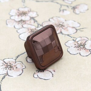May include: A square-shaped cabinet knob with a brown, metallic finish. The knob has a textured, diamond-like pattern on the surface.
