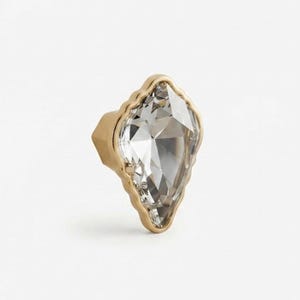 Peut inclure: Un bouton décoratif doté d'un cristal transparent à facettes en forme de cerf-volant, serti dans un cadre en métal doré festonné. La pièce présente une base pointue et une surface lisse et réfléchissante au design géométrique.