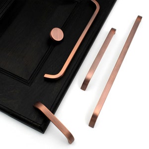 May include: Four copper cabinet pulls in different styles.  The pulls are on a white background.  One pull is a round knob, and the other three are bar pulls.  Two of the bar pulls are straight, and one is curved.