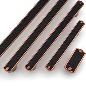 Könnte beinhalten: Eine Sammlung von bronzefarbenen Schrankgriffen mit einem schwarzen, strukturierten Mittelteil. Die Griffe sind unterschiedlich groß, mit einem rechteckigen Knauf am Ende des kleinsten Griffs. Die Hardware hat ein gehämmertes Aussehen.