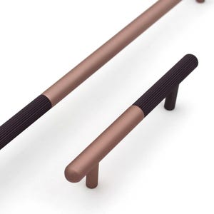 May include: Close-up of two cabinet pulls. The pulls are a muted rose gold color with a dark brown, ribbed grip. One pull is long and straight, while the other is shorter with a curved handle. Hardware for cabinets.