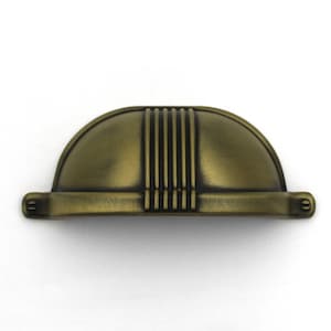 May include: A semi-circular cup pull handle in an antique brass finish with a brushed texture. The hardware features a central vertical ribbed design and two small circular mounting points on each side, each containing a single visible screw head.
