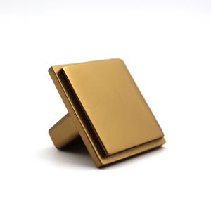 Peut inclure: Un bouton de meuble carré avec une finition dorée mate. Le bouton présente une face carrée plate et surélevée avec un bord étagé, créant un aspect superposé. La base du bouton est inclinée, montrant une couleur dorée métallique uniforme.