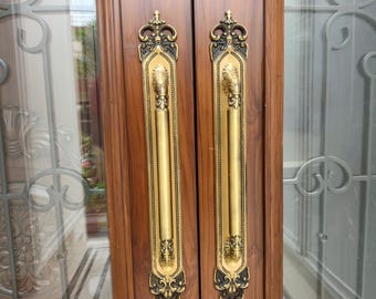 May include: A pair of long, vertical brass door handles with intricate, ornate detailing at the top and bottom, mounted on a polished brown wooden door frame. The handles feature a smooth central grip and decorative scrollwork accents.