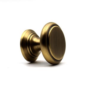 Peut inclure: Un bouton de meuble rond avec une finition en laiton antique brossé. La quincaillerie présente une face circulaire lisse et légèrement bombée avec une base étagée, affichant une patine dorée métallique et brun foncé sur toute la surface.