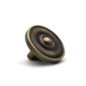 May include: A round cabinet knob with an antique brass finish, featuring a central dome surrounded by a series of radiating, fine grooves that extend to the outer edge of the circular face, creating a textured and detailed metallic appearance.