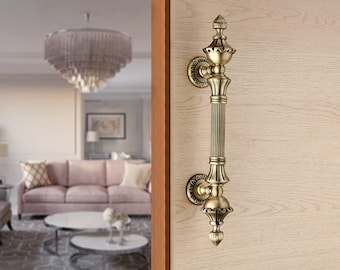 Puede incluir: Una manilla de puerta dorada con un diseño ornamentado. La manilla es larga y tiene un remate decorativo en la parte superior. La manilla está montada en una puerta de madera.