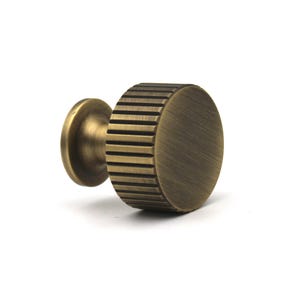 Op de afbeelding: Een cilindrische meubelknop met een geborstelde antiek messing afwerking. De knop heeft een plat, rond voorvlak en een zijkant met verticale ribbels. Het bevat een kleine, gladde, ronde basis die verbonden is met het hoofdgedeelte.
