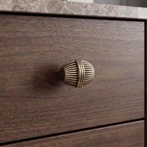 Puede incluir: Un pomo de mueble de forma ovalada con un acabado de latón metálico y una serie de estrías verticales. El pomo está montado en un frente de cajón de madera de color marrón oscuro que presenta un patrón de veta horizontal y textura mate.