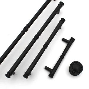May include: A collection of matte black cabinet hardware including three elongated bar pulls of varying lengths and one circular knob. Each pull features a central decorative ribbed section and flared ends, while the knob has a beaded border.