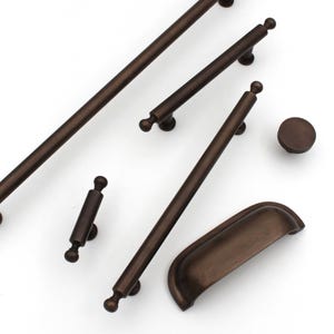 May include: A collection of bronze-colored cabinet hardware, including various sizes of handles and a knob. The handles are cylindrical with rounded ends, and one is a cup pull. The hardware is arranged on a white background.