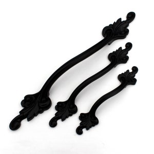 May include: Three black cabinet handles with ornate, leaf-like designs at each end. The handles vary in size, with the longest measuring approximately 30 cm in length. The handles are made of a matte black material.