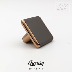 Puede incluir: Un tirador de armario cuadrado con una cara gris oscuro y un borde y una base dorados. La palabra "Luxury" está escrita en cursiva, con "By ABVIN" debajo. Un logotipo está en la esquina superior derecha.