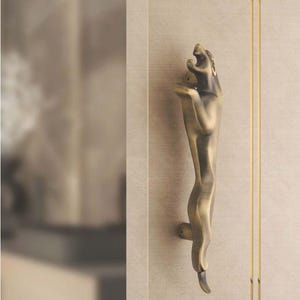 May include: Bronze-toned door handle with a stylized animal figure design. The handle is mounted on a light-colored wall with vertical gold accents. The animal figure is in a standing pose.