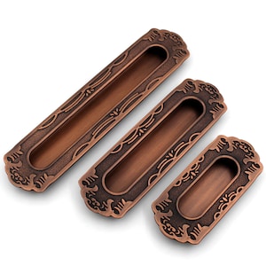 Pode incluir: Três puxadores de gaveta em tom cobre antigo com designs ornamentados. Cada puxador tem uma forma retangular com um recorte no centro. Os puxadores estão dispostos em ordem decrescente de tamanho.