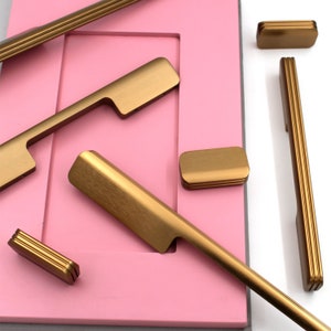 Könnte beinhalten: Sortiment an goldenen Schrankbeschlägen, Griffen und Knöpfen auf rosa Untergrund. Die Beschläge umfassen einen langen Stangen-Griff, einen kürzeren Stangen-Griff, einen kleinen Knopf und einen größeren Knopf.