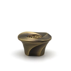Könnte beinhalten: Ein bronzefarbener Schrankknopf mit einem dekorativen, wirbelnden Design. Der Knopf hat einen ausgestellten Sockel und ein zentrales, kreisförmiges Element mit einem durchbrochenen Muster. Die Gesamtform erinnert an eine stilisierte Blume oder eine kleine, elegante Skulptur.