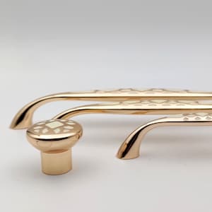 Puede incluir: Juego de herrajes dorados para armarios con un pomo y dos tiradores curvos, todos con un patrón geométrico crema. Los herrajes están diseñados para muebles y tienen un acabado pulido.
