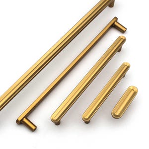 May include: A collection of gold-colored cabinet pulls and handles in various sizes. The hardware features a grooved design and a warm, metallic finish. The pulls are arranged diagonally against a white background, showcasing their different lengths.