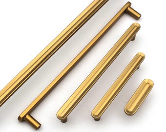 May include: A collection of gold-colored cabinet pulls and handles in various sizes. The hardware features a grooved design and a warm, metallic finish. The pulls are arranged diagonally against a white background, showcasing their different lengths.