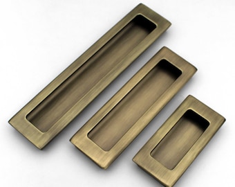 May include: Three antique brass-toned recessed drawer pulls in varying sizes. Each pull has a rectangular shape with a recessed finger pull area. The pulls are arranged diagonally on a white background, showcasing their aged finish.