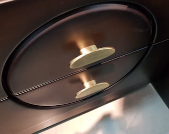 Puede incluir: Primer plano de una cómoda marrón oscuro con dos cajones. Cada cajón tiene un tirador dorado de forma ovalada. La cómoda tiene un acabado liso y brillante, y los tiradores reflejan la luz.