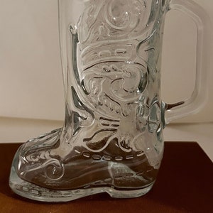 May include: Clear glass beer stein shaped like a cowboy boot. The glass has embossed decorative scrollwork and a handle. The boot-shaped base transitions into a cylindrical upper portion. The glass is transparent, allowing visibility of the contents.