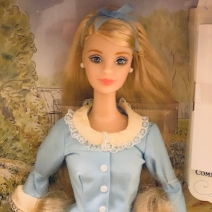 以下が含まれることがあります： 白い襟と袖口が付いた水色のドレスを着たブロンドのドール。ドレスはチェック柄のスカートで、前面にボタンが付いています。ドールは青い目を持ち、髪にはお揃いの青いリボンを付けています。ドールは風景の背景がある箱に入っています。