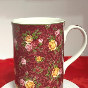May include: A ceramic mug with a white handle and rim. The mug features a burgundy background with a pattern of yellow, pink, and red roses with green leaves. The mug is sitting on a white surface.