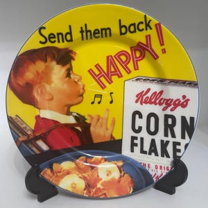 Peut inclure: Assiette décorative vintage avec un fond jaune et les mots "Send them back HAPPY!" en noir et rouge. L'assiette représente un garçon et une boîte de Kellogg's Corn Flakes. Un petit plat avec des céréales est devant.