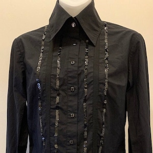 May include: A black long-sleeved button-down shirt featuring a structured collar and a central button placket. Two vertical rows of black sequins run down the front of the shirt, flanking the button closure, adding a textured detail to the fabric.