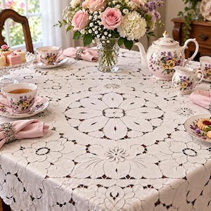 May include: A white tablecloth featuring intricate cutwork embroidery with large floral motifs and scalloped edges. The table is set with a floral porcelain tea set, pink napkins in silver napkin rings, and a glass vase filled with pink roses.