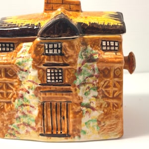 Peut inclure: Un pot à thé en céramique en forme de cottage avec un toit brun, des murs beige et des vignes vertes peintes. La structure présente une porte centrale, plusieurs fenêtres à carreaux et une cheminée sur le dessus. L'extérieur est brillant.