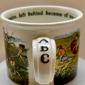 May include: A cream-coloured ceramic mug featuring colourful illustrations of small winged figures and birds in a grassy landscape. The handle is embossed with the letters A, B, and C in black. Text is printed along the inner rim of the mug.