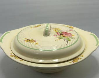 May include: Cream-colored ceramic serving dish with a lid. The lid features a floral design with pink roses and yellow flowers, accented by green stripes and a handle. The dish has handles on either side.