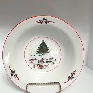 May include: White ceramic bowl with a red rim and a Christmas scene. The bowl features a central image of a decorated Christmas tree, houses, and a sleigh. Holly and ribbon accents are placed around the rim.