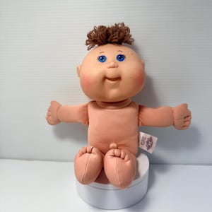Peut inclure: Une poupée au corps souple avec un visage rond aux tons pêche, des yeux bleu vif et une petite touffe de cheveux en laine brune sur le dessus de la tête. La poupée a des joues roses, un léger sourire et une étiquette blanche avec CPX imprimé.