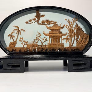 Peut inclure: Un objet décoratif ovale présentant une sculpture détaillée en liège brun clair représentant une pagode, des arbres et des formations rocheuses dans un cadre noir. La scène est placée dans une enceinte ovale vitrée sur un socle en bois noir.